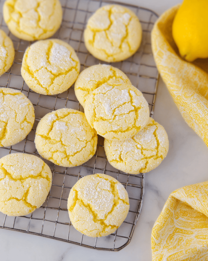 Lemon Cake Mix Cookies: Easy, Soft, and Delicious! 14 Stack of soft, yellow lemon cake mix cookies dusted with powdered sugar, arranged on a white plate with a blurred floral background.